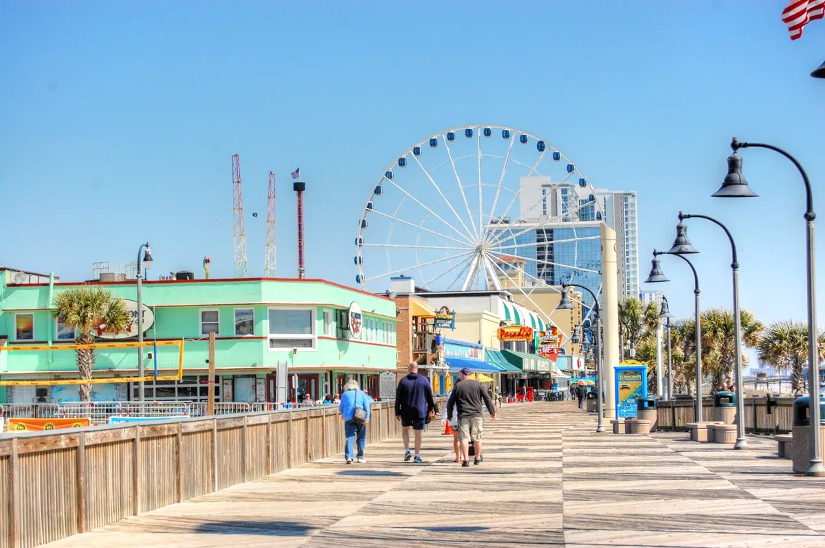 A lively Myrtle Beach boardwalk scene showcases the iconic ferris wheel, colorful buildings, palm trees, and visitors walking past shops and eateries. The joyful mood is enhanced by people enjoying rides and sampling local food.