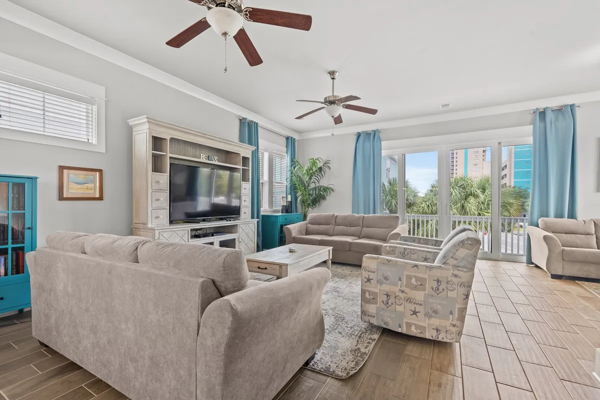 A spacious living area featuring a large, comfortable seating arrangement, abundant natural light streaming through oversized windows, and a stylish entertainment unit with a flat-screen TV. The décor is contemporary with coastal influences, creating an inviting and relaxing atmosphere for gatherings.