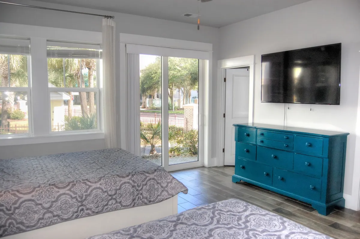 A welcoming bedroom with two beds featuring patterned bedding, a vibrant blue dresser, large windows with views outdoors, and a flat-screen TV. The space feels both comfortable and flexible, accommodating families or groups and providing entertainment and natural light.