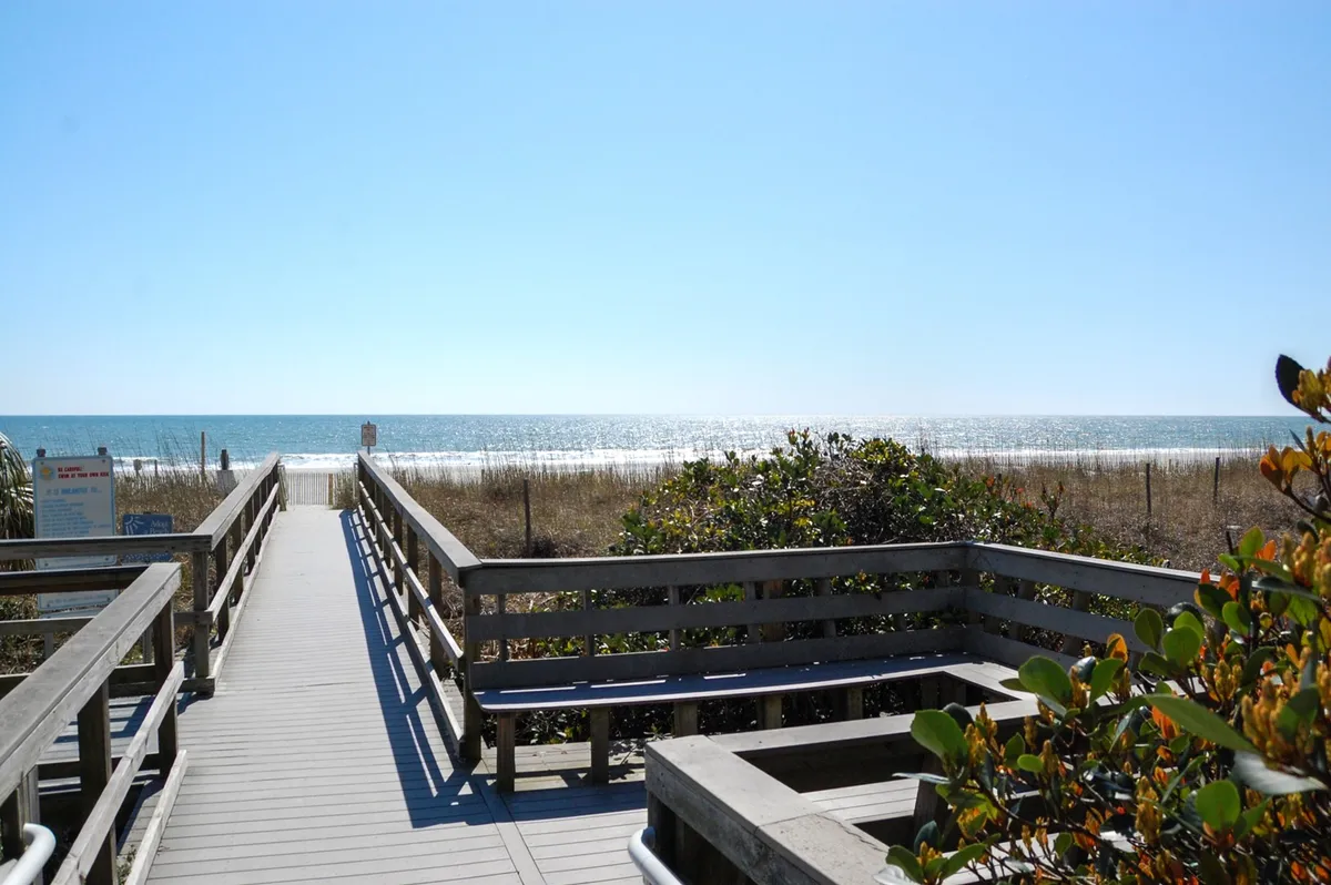 A wooden walkway extends over grassy dunes, leading directly to the sandy beach with a wide ocean view. The scene is bathed in natural light, highlighting the easy access from the property to the shoreline.
