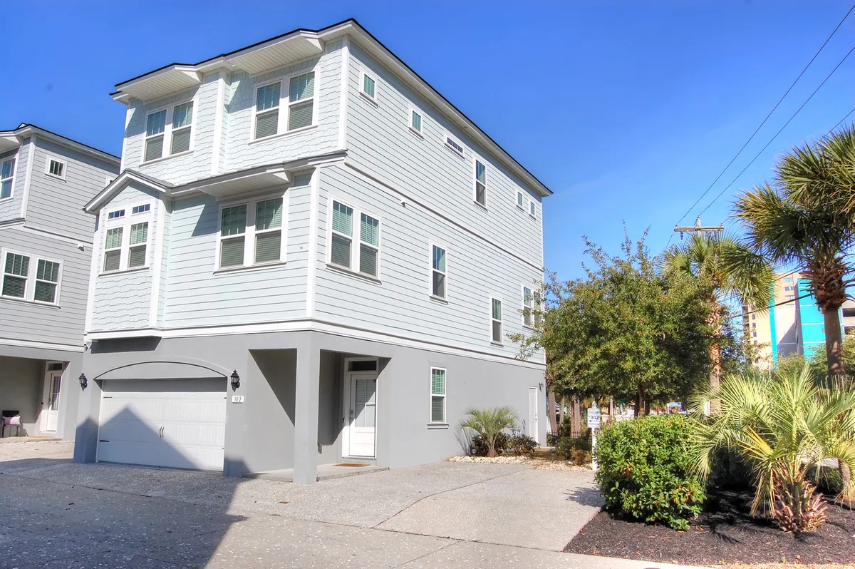 The image features a bright, welcoming two-story vacation home surrounded by swaying palm trees, with large windows letting in sunlight and a spacious driveway out front. The inviting porch hints at sunny afternoons and warm evenings spent outdoors with family and friends, all just moments from the beach.