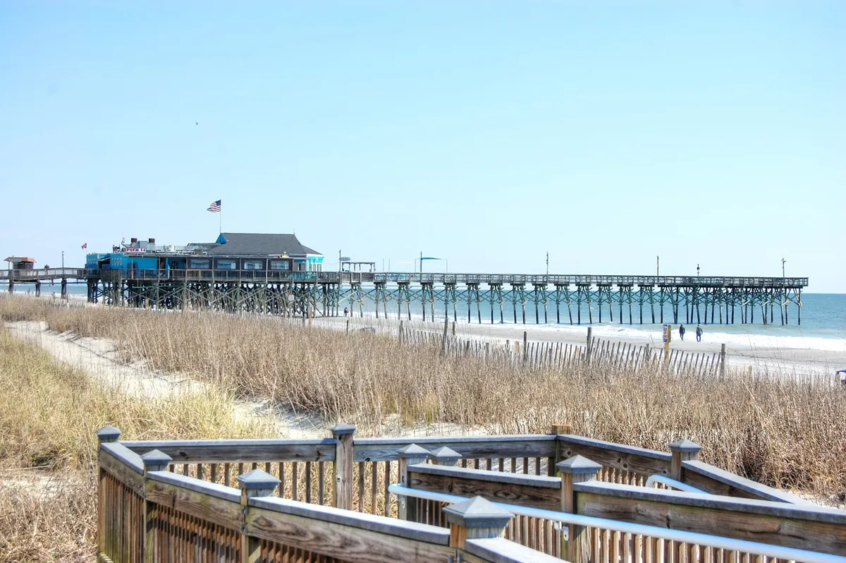 A scenic shot featuring a classic pier stretching out over the sandy beach, bordered by dune grass and gentle waves. The scene captures people strolling along the pier, kids playing in the sand, and the expansive shoreline—evoking the quintessential family beach day described in the section.