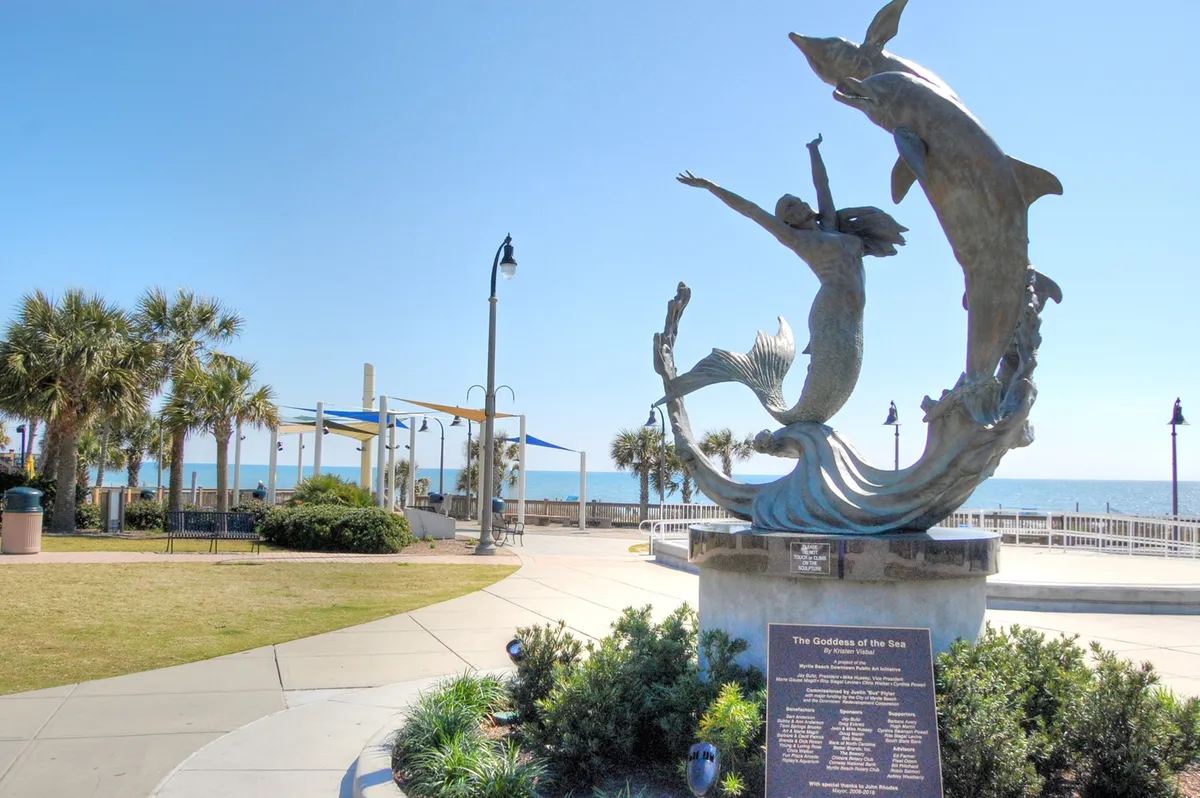 A beautiful outdoor scene featuring the iconic Goddess of the Sea statue, graceful dolphins, and palm trees, with the ocean and Myrtle Beach Boardwalk in the background. The area is lively yet peaceful, capturing the essence of local attractions and the natural beauty that retirees and snowbirds can explore nearby.